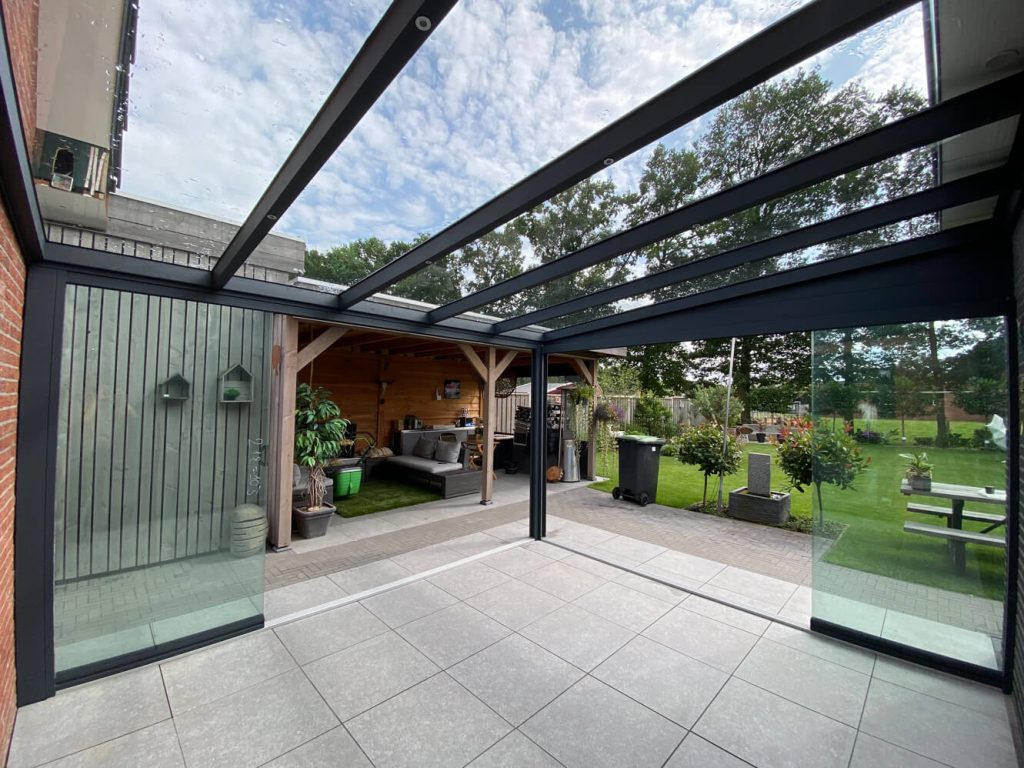 Moderne Aluminium Terrassenüberdachung mit offenem Glasdach und Glasschiebewänden, mit Blick auf einen liebevoll gestalteten Garten.