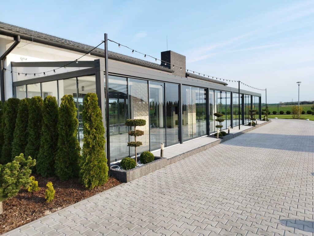 Große gläserne Überdachung mit schwarzem Metallrahmen bei einer Gastronomiestätte mit geräumiger Terrasse und stimmungsvoller Beleuchtung.