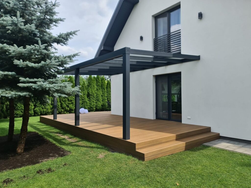Moderne Aluminium Terrassenüberdachung mit schwarzem Metallrahmen und Holzboden, umgeben von einem grünen Garten und einer großen Kiefer.