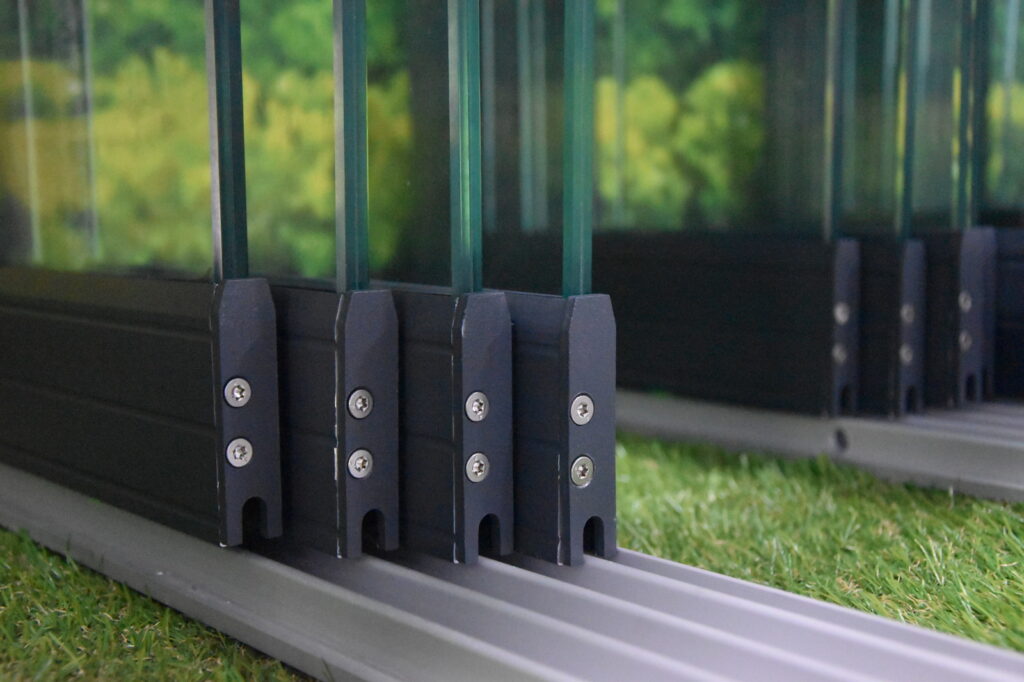 Nahaufnahme eines Glasschiebesystems mit Metallschienen auf einer rasenähnlichen Unterlage, mit grünem Spiegelbild in den Glasflächen.