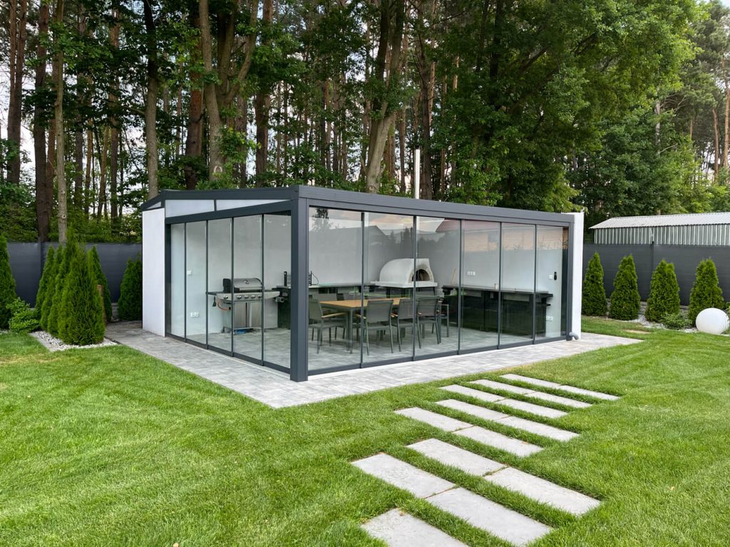 Freistehender gläserner Gartenpavillon mit Aluminiumrahmen, eingerichtet als Außenküche und Essbereich, umgeben von einem grünen Garten und Bäumen.