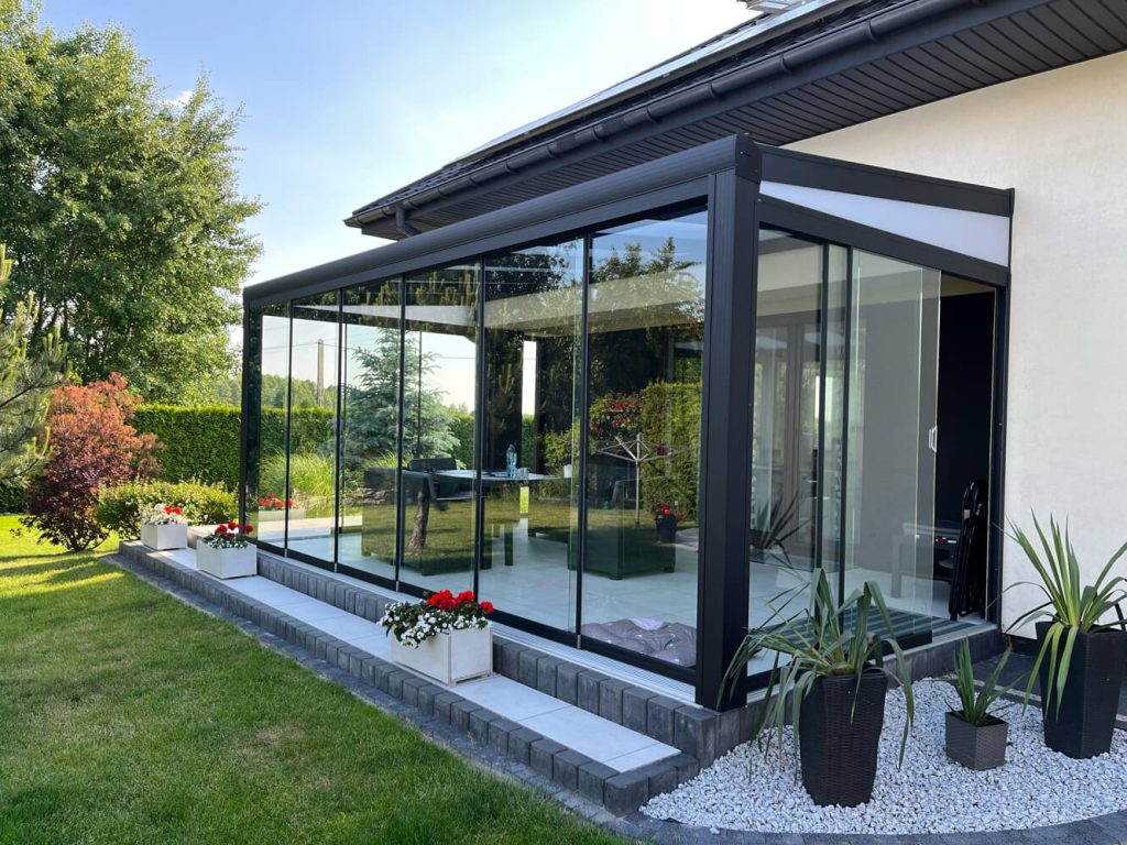 Stilvoller Wintergarten mit Glasschiebewänden und modernem Design, angebaut an ein Haus mit erhöhtem Terrassenbereich, umgeben von einem gepflegten grünen Garten.
