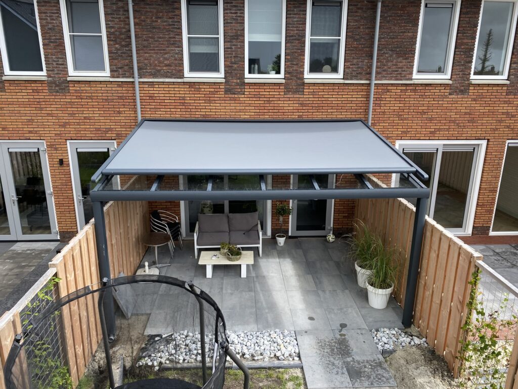 Terrassenüberdachung mit Sonnenschutz und Glasschiebewänden, eingerichtet mit Stühlen und Pflanzen, angrenzend an ein Reihenhaus, umgeben von Zäunen.