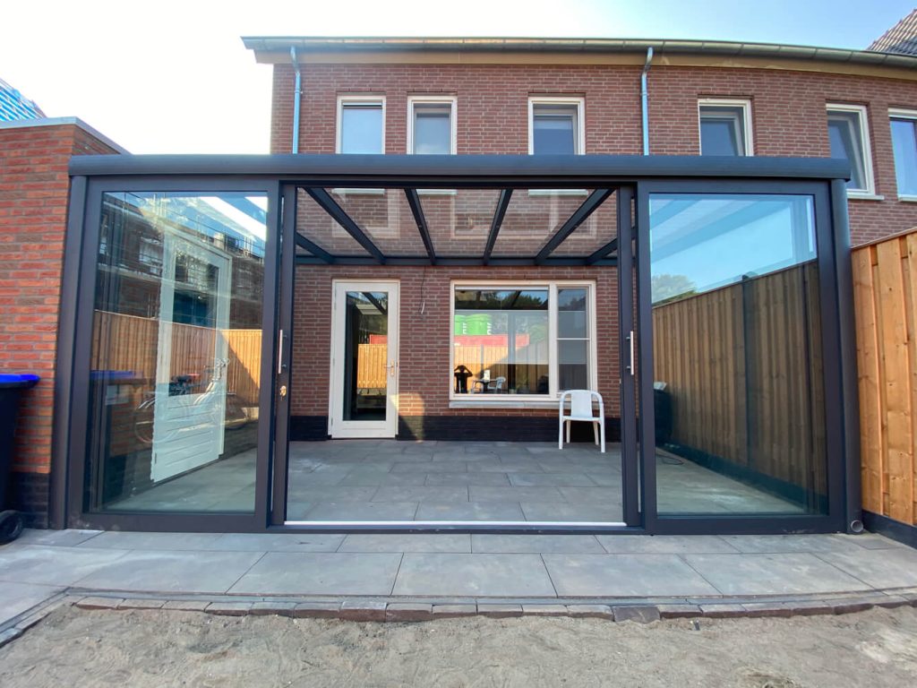 Schlichter Wintergarten mit Glasdach und Glasschiebewänden, angebaut an ein modernes Wohnhaus, mit Fliesenboden und Holzzäunen im Garten.