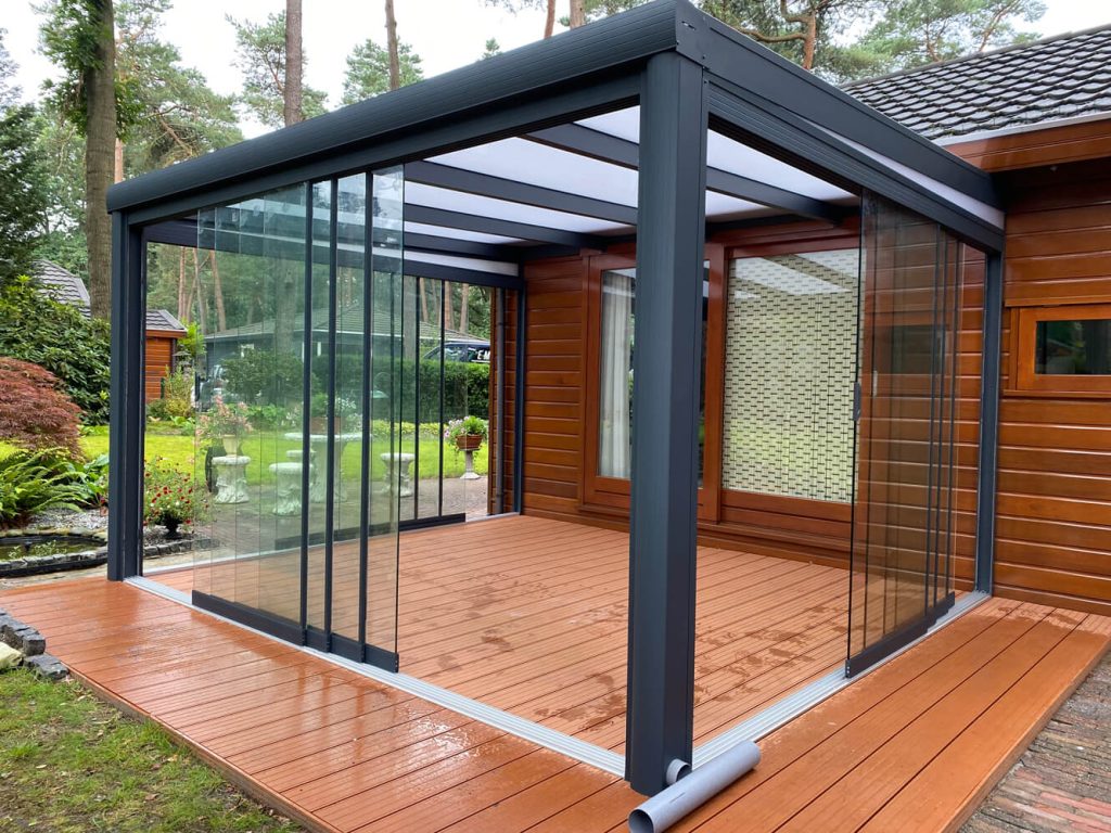 Kleine Aluminium Terrassenüberdachung mit Glasschiebewänden und integriertem Sonnenschutz, angebaut an ein Ziegelhaus.