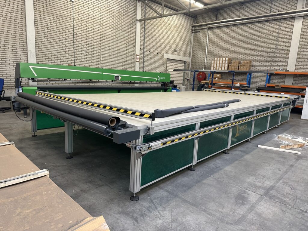 Große Industrieschneidemaschine in einem Lager, verwendet zur Verarbeitung von Sonnenschutzstoffen, mit grünem Gehäuse und gelbem Warnstreifen.
