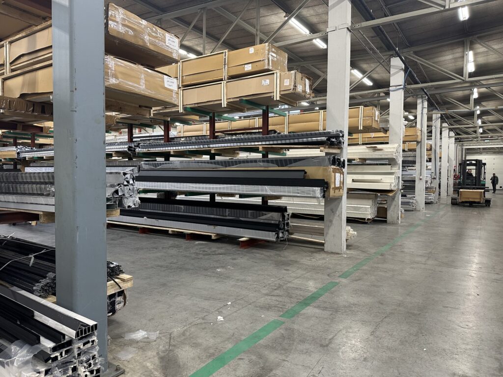 Übersicht eines Lagerraums mit Metallträgern und Aluminiumteilen auf Holzpaletten unter Leuchtstofflampen und mit hoher Decke.