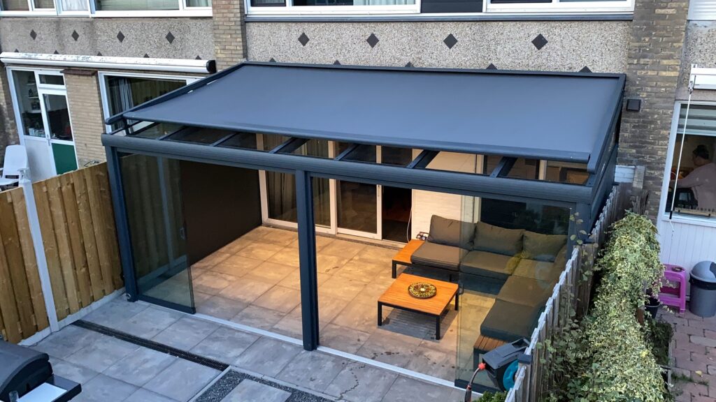 Moderne Aluminium Terrassenüberdachung mit Sonnenschutz und Glasschiebewänden (10mm gehärtetes Glas), ausgestattet mit einer Sitzecke, grauen Fliesen, Holzsichtschutz und Grünfläche.