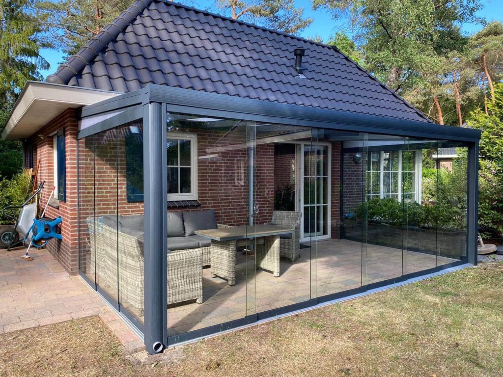 Gemütlicher Wintergarten mit Glasschiebewänden und moderner Ausführung, angebaut an ein Ziegelhaus, eingerichtet mit einer Loungegarnitur in grüner Umgebung.