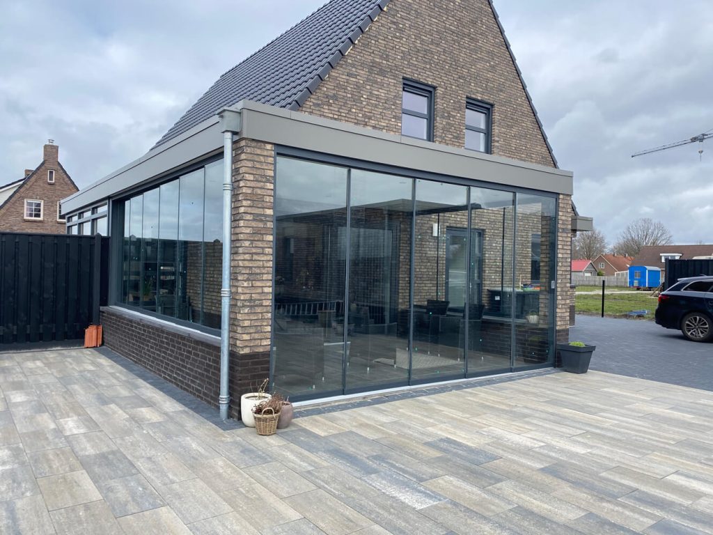 Moderner Wintergarten mit Glasschiebewänden und klarer Ausführung, angebaut an ein Steinhaus mit großzügiger gefliester Terrasse und freiem Ausblick.