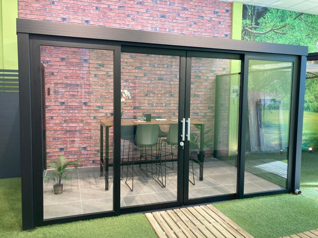 Moderne Aluminium Terrassenüberdachung mit Glasschiebewänden und schwarzem Metallrahmen, eingerichtet mit einem Hochtisch und Stühlen an einer Ziegelwand mit grünen Akzenten.