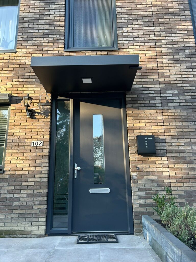 Schwarze Aluminium Terrassenüberdachung über einer Haustür eines Hauses mit Ziegelfassade, mit integriertem Lichtpaneel.