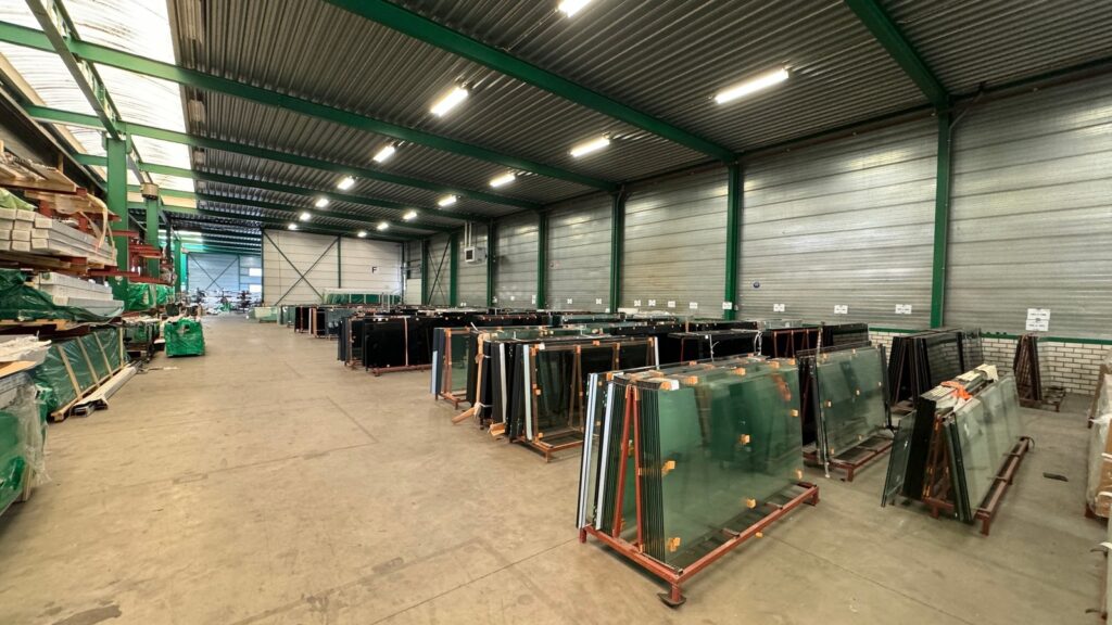 Übersicht eines Lagerbereichs mit Glasplatten in Metallregalen, mit grüner Stahlkonstruktion und Arbeitsbereichen.