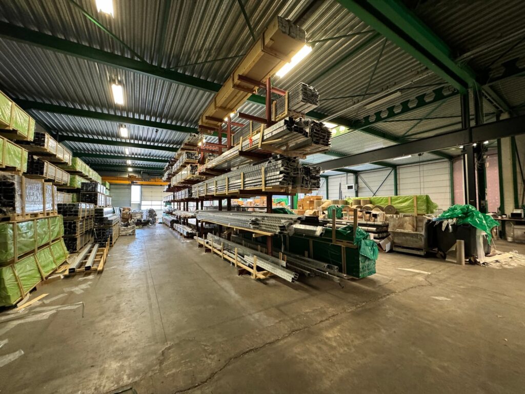 Lager mit gestapelten Aluminiumprofilen und anderen Baumaterialien, in industriellen Regalen unter einem Metalldach gelagert.