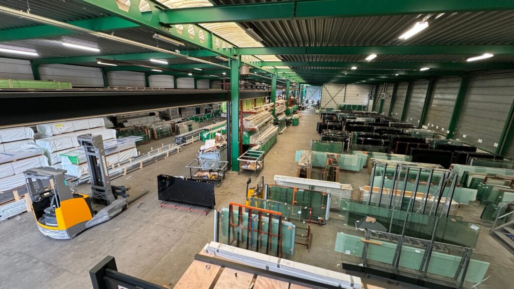 Übersicht über ein großes Lager mit Stahlkonstruktionen, Glasplatten und Baumaterialien, inklusive Gabelstaplern und Arbeitsstationen.
